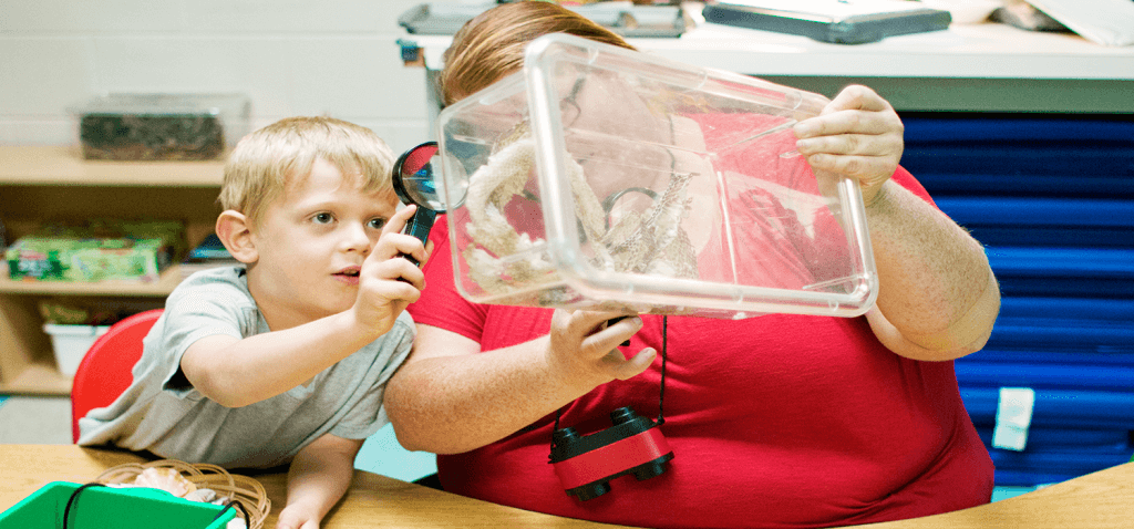 A boy is holding a magnifying glass to a clear box which a teacher is holding. Inside the clear box appears to be snake skins.