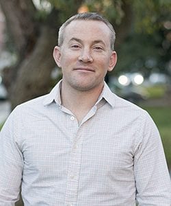 A photo of Aaron standing outside in front of a tree smiling at the camera.