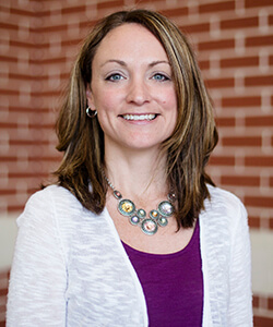 A photo of Hallie standing outside in front of a brick wall smiling at the camera.