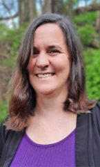 Photo of a woman smiling at the camera wearing a purple shirt with a black jacked standing in front of green trees