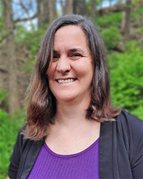 Photo of a woman smiling at the camera wearing a purple shirt with a black jacked standing in front of green trees