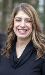 Photo of a woman who is smiling wearing a black sweater stanging in front of a black fence and a garden.