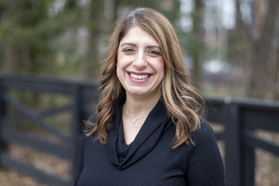 Photo of a woman who is smiling wearing a black sweater stanging in front of a black fence and a garden.