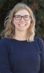 photo of a woman with blonde hair and glasses wearing a navy sweater standing in front of a green tree