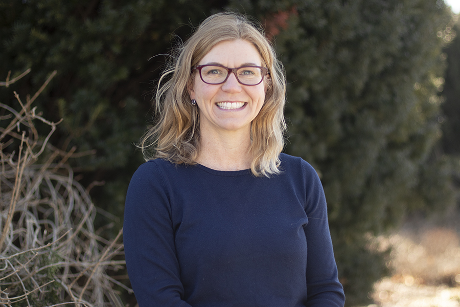 photo of a woman with blonde hair and glasses wearing a navy sweater standing in front of a green tree