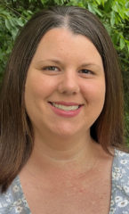 headshot of a lady with long brown hair wearing a fblue floral shirt, smiling while standing in front of foilage.