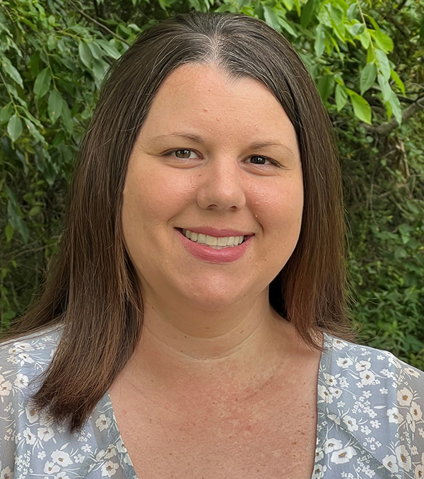 headshot of a lady with long brown hair wearing a fblue floral shirt, smiling while standing in front of foilage.