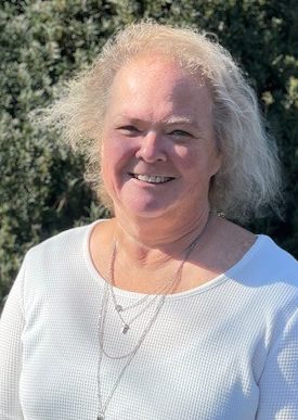 Woman in a white shirt smiling and standing in front of green foliage.