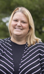 Woman with blonde hair, smiling, wearing a black and white striped sweater standing in front of trees.