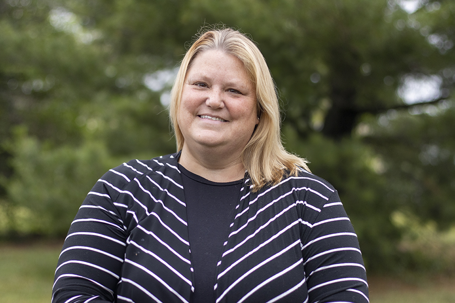 Woman with blonde hair, smiling, wearing a black and white striped sweater standing in front of trees.