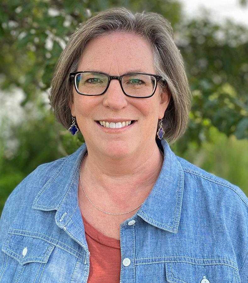 Woman with short light brown hair wearing a orange shirt with a denim jacket