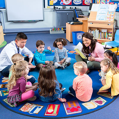 Group of nursery children sitting on the floor in their classroom with their teachers. The female teacher is reading from a book.