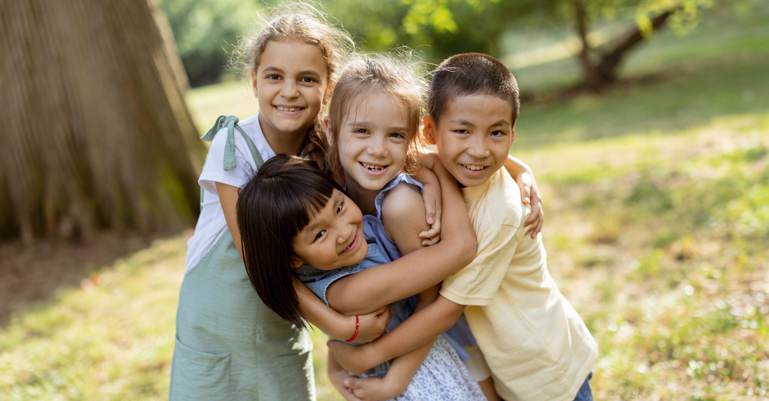 Four young children hugging each other in a par setting.