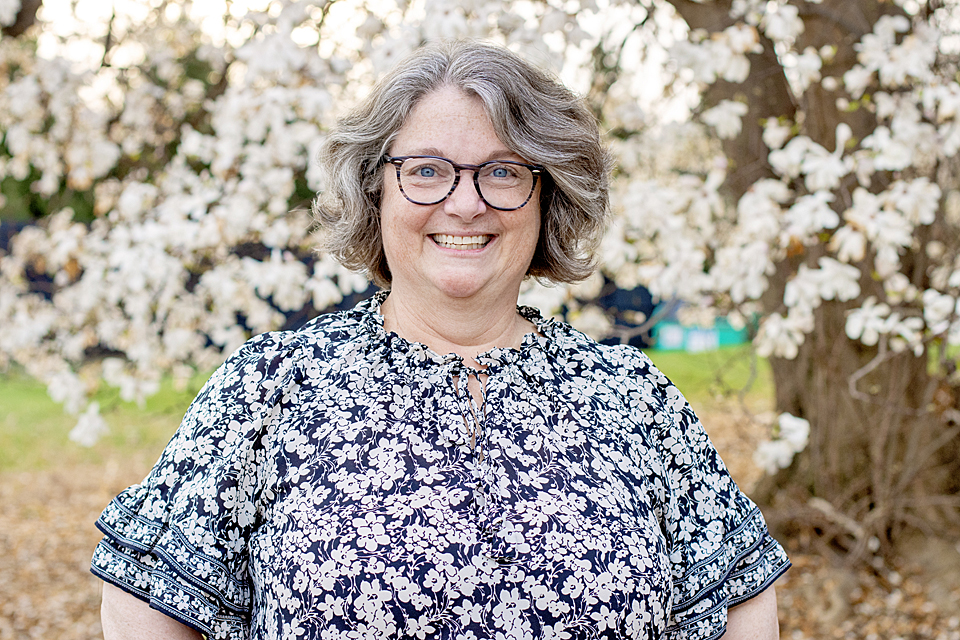 Christine Hausman is sitting in a portrait studio facing the camera. She is a middle aged white woman with greying short hair. Christine is wearing a black sweater.