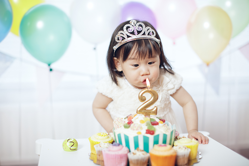 Yong Asian child wearing a silver crown blowing out a number 2 candle on a birthday cake.