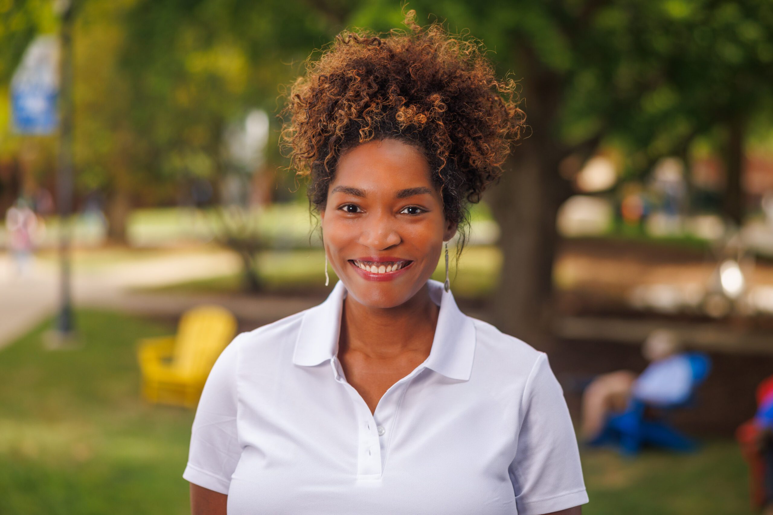 African American woman smiling at the camera wearing a white polo shirt with her curly light brown hair in a high ponytail on her head.