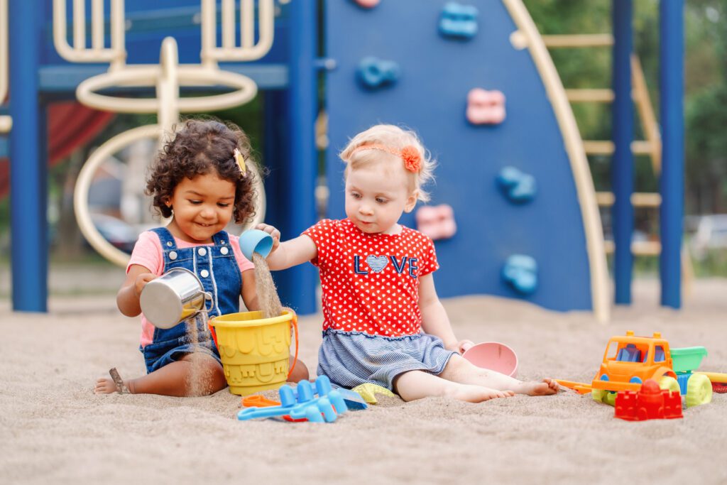 Two toddler girls play with a bucket of sand outdoors.