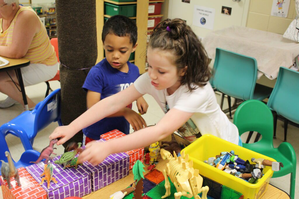 Two children playing with dinosaurs and other toys at a table.