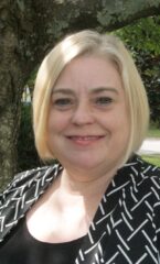 White woman with short blonde hair wearing a black and white patterned top smiles for the camera.