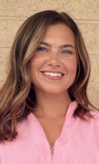 A woman with medium length, light brown hair who is wearing a pink blouse in front of a tan brick wall.