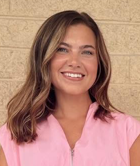 A woman with medium length, light brown hair who is wearing a pink blouse in front of a tan brick wall.