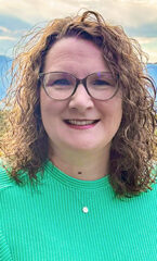 Woman with shoulder length, curly brown hair wearing glasses and a teal blouse.