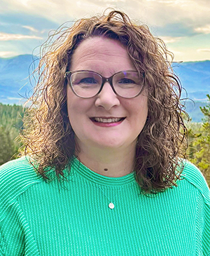 Woman with shoulder length, curly brown hair wearing glasses and a teal blouse.